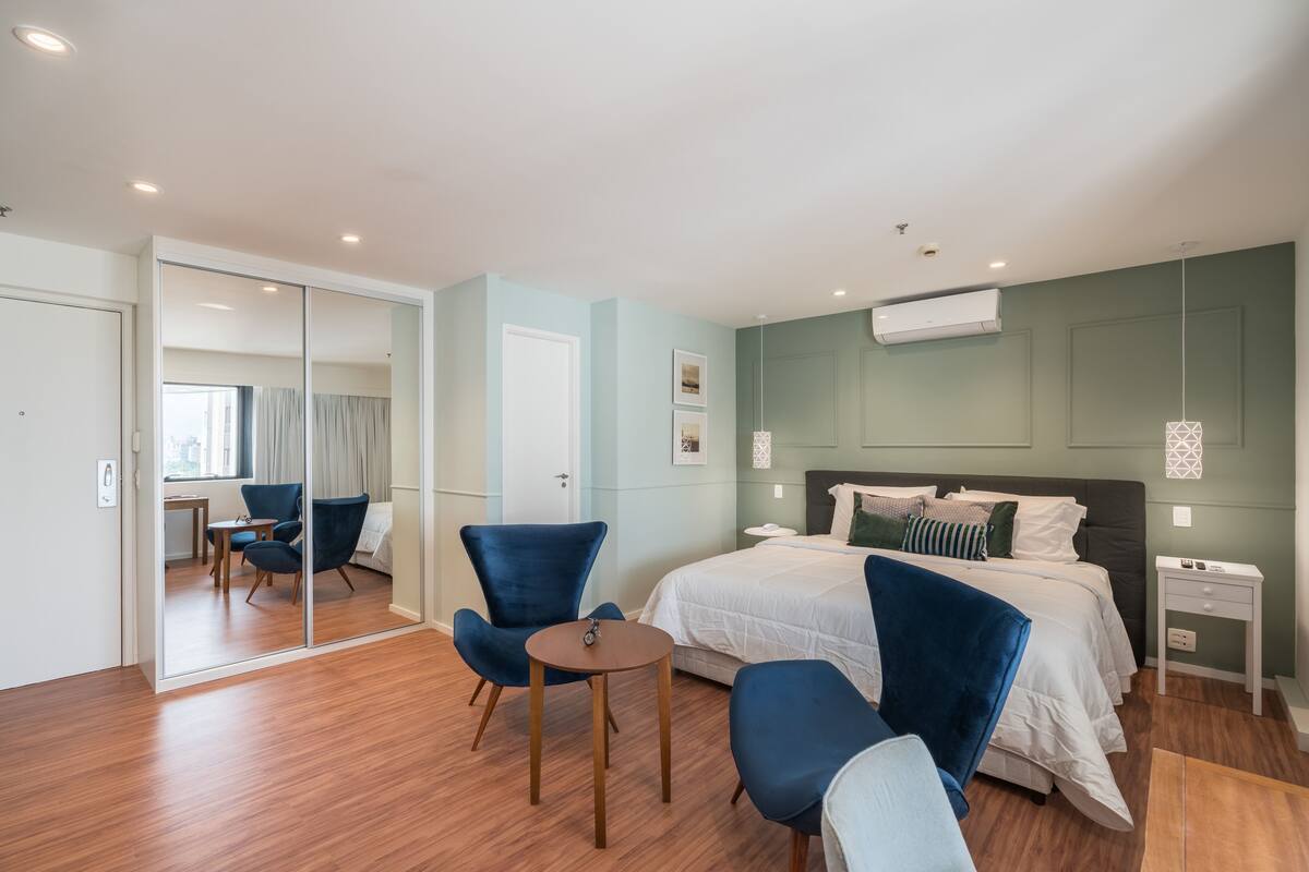 A modern hotel room with a large bed, two blue armchairs, a small round table, a bedside table, and a mirror.