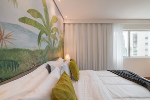 Habitación de hotel con una cama grande, ropa de cama blanca, almohadas verdes, una mesita de noche con lámpara, un mural de plantas tropicales y una ventana con cortinas.