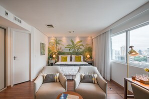 Een hotelkamer met een groot bed, twee fauteuils, een bureau met lamp en uitzicht op de stad.