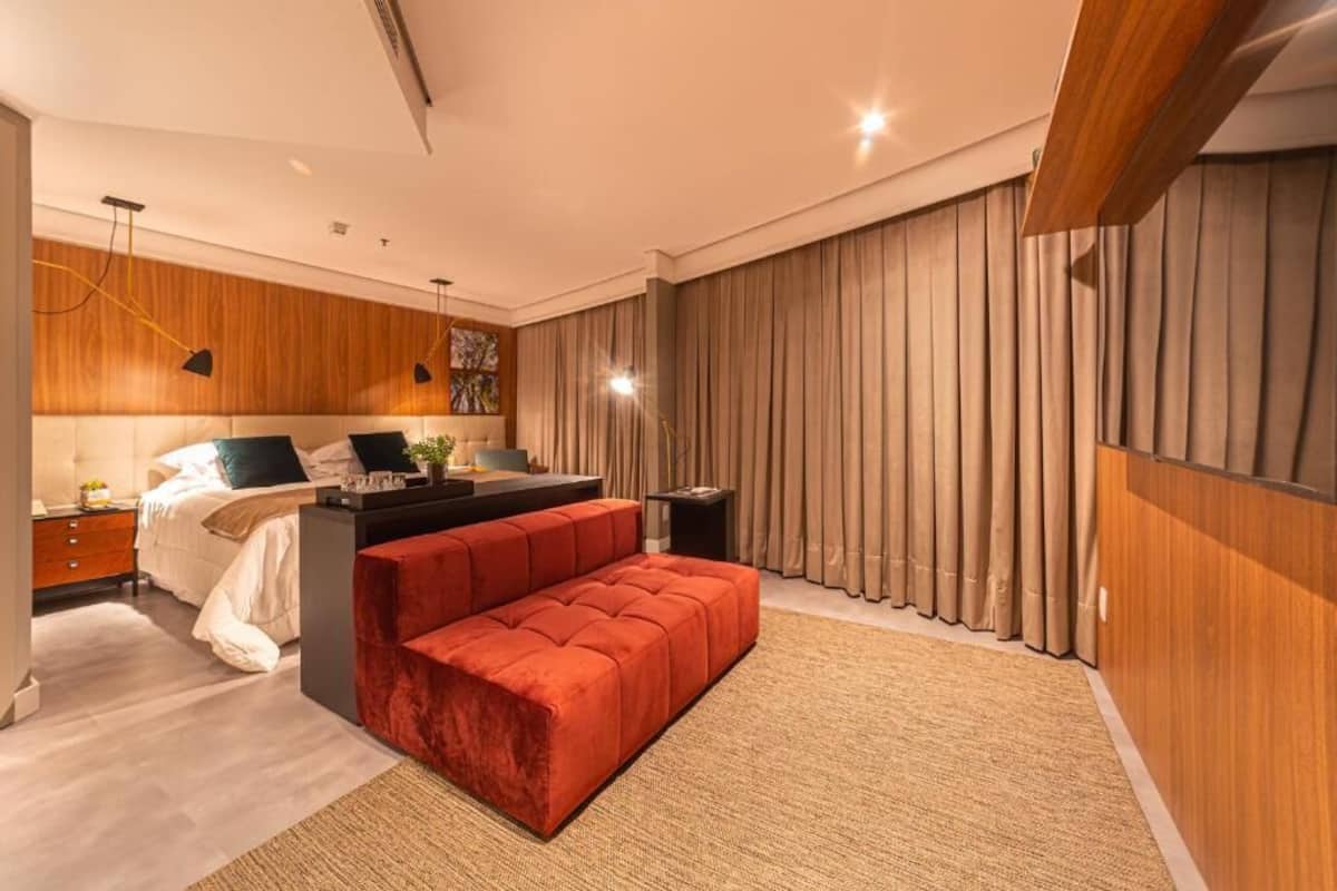 A modern bedroom with a bed, a red ottoman, a desk, and a large window with curtains.