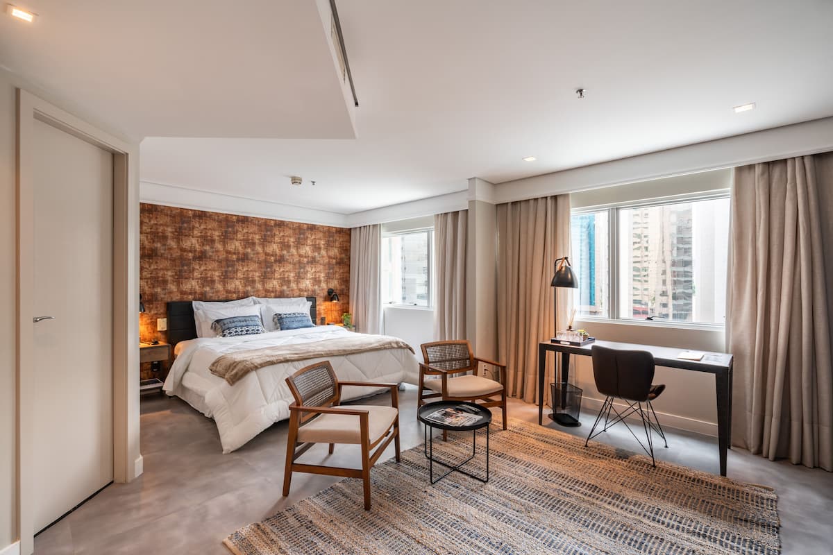 A modern hotel room with a large bed, two chairs, a desk, and a view of the city.