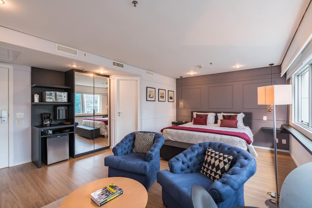 A modern hotel room with a bed, two blue armchairs, a small coffee table, a desk with a computer, and a large window.