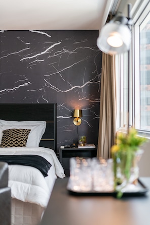 A modern bedroom with a black marble wall, a bed with white bedding, a gold lamp, and a window with curtains.