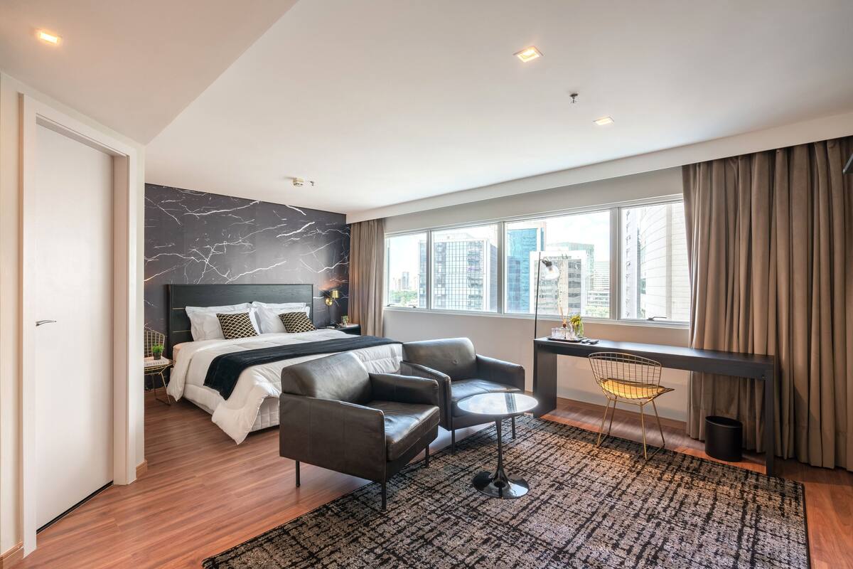A modern hotel room with a large bed, a sofa, a small table, and a view of the city through large windows.