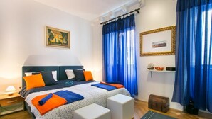 Un dormitorio con una cama, dos otomanas blancas, una mesita de noche, una lámpara, una ventana con cortinas azules, un cuadro enmarcado en la pared y un pequeño estante con objetos decorativos.