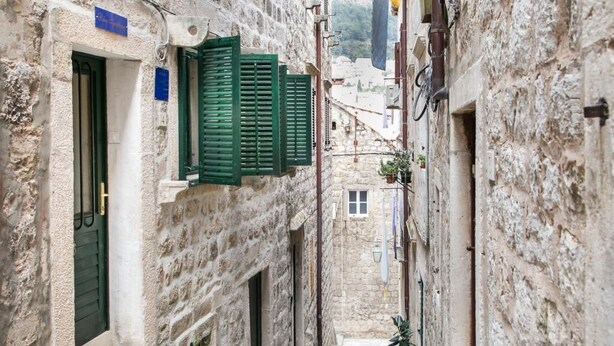 Un estrecho callejón de piedra con una escalera que lleva a una puerta, flanqueado por edificios con contraventanas verdes y plantas en macetas.