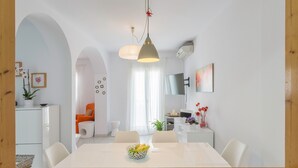 Un espace de salle à manger moderne avec une table blanche, des chaises et un saladier rempli de fruits. On y trouve deux lampes suspendues, un climatiseur blanc et un téléviseur fixé au mur.
