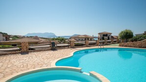 A swimming pool with stone tiles, a stone wall, and a view of mountains in the background. - Holiday Home 'Sardinia Sea View Pool & Spa' with Pool and Hot Tub (pittulongu)