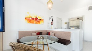 Uma sala de estar moderna com uma mesa de vidro, cadeiras de vime, um sofá com almofadas, uma cozinha com armários brancos e um quadro na parede.