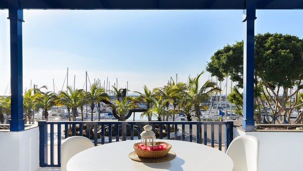 Mesa redonda com cadeiras, um candeeiro e uma cesta de frutas numa varanda com vista para uma marina com barcos e palmeiras.