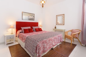 Une chambre à coucher bien rangée, avec une tête de lit rouge, un lit simple avec une literie rouge et blanche, une chaise en osier, une table de chevet avec une lampe et un tableau encadré au mur.