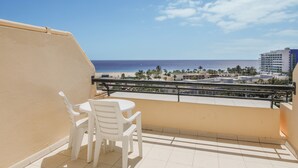 Un balcón con vista a la playa y al océano, una mesa y sillas blancas, y una barandilla.