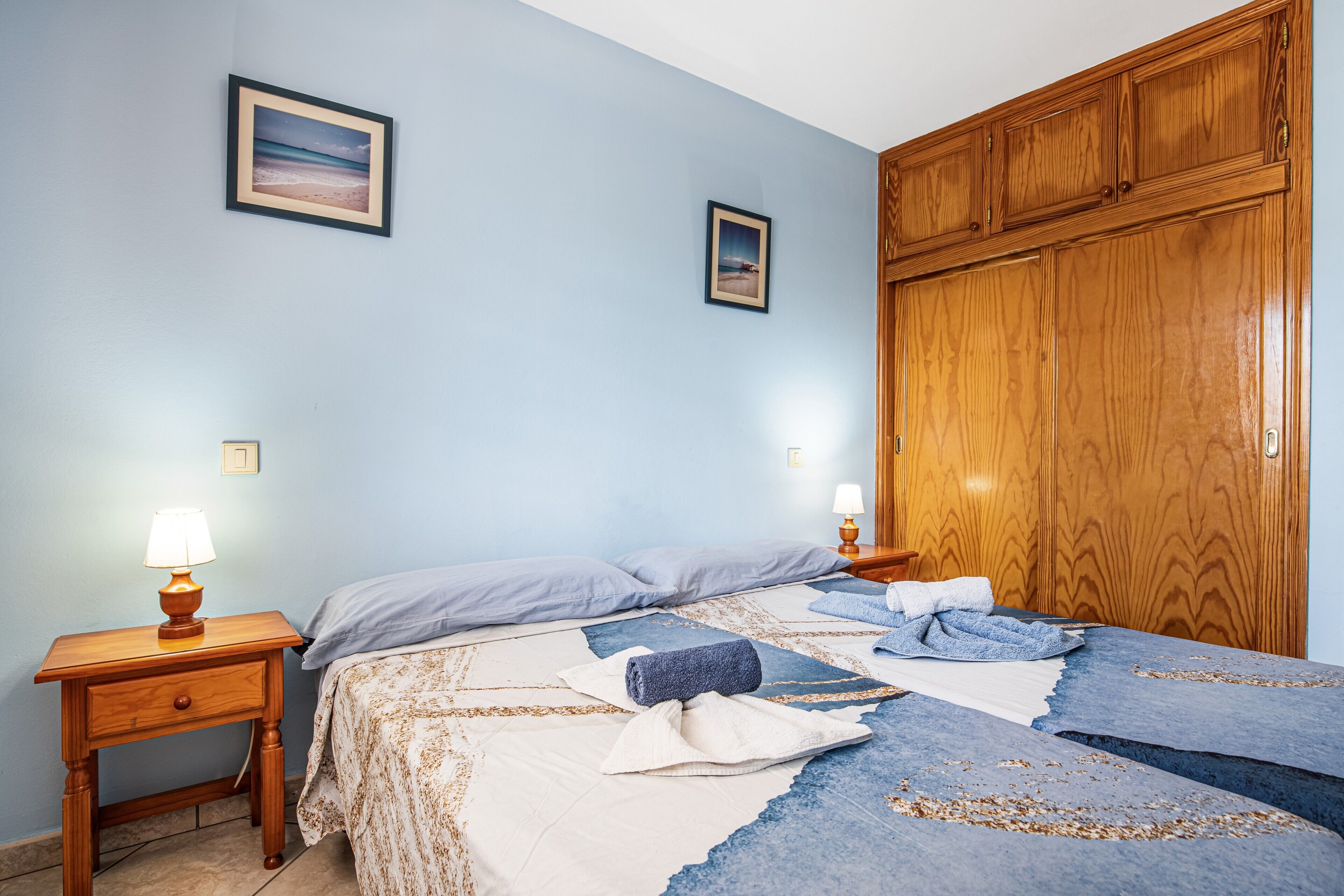 Une chambre à coucher comprenant un lit, une armoire en bois, deux tables de chevet avec des lampes et deux tableaux encadrés accrochés au mur.