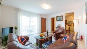A modern living room with a brown leather sofa, a coffee table, and a TV. There is a large window with curtains, a door leading to another room, and a small table with a vase and a plant. - Feel Good Apartment (Dubrovnik)
