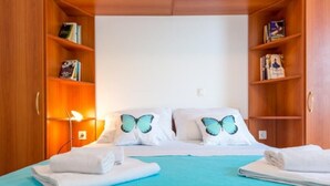 A hotel room with a bed, two pillows with butterfly designs, a blue bedspread, and a wooden wardrobe with books and a lamp. - Feel Good Apartment (Dubrovnik)