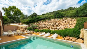 Ein Schwimmbecken mit Liegestühlen und Sonnenschirmen, umgeben von einer Steinmauer und viel Grün.
