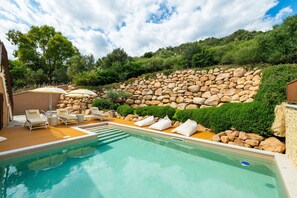 A swimming pool with lounge chairs and umbrellas, surrounded by a stone wall and greenery.