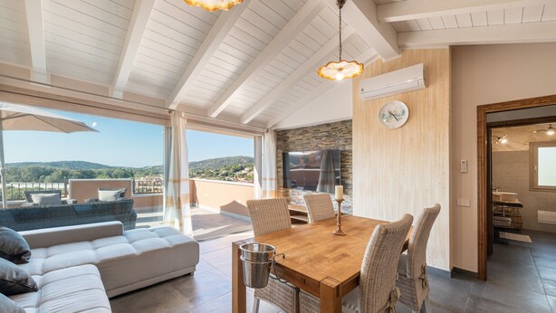 Ein geräumiger Wohnbereich mit einem Holzessttisch, Korbstühlen, einer Couch und Blick ins Freie.