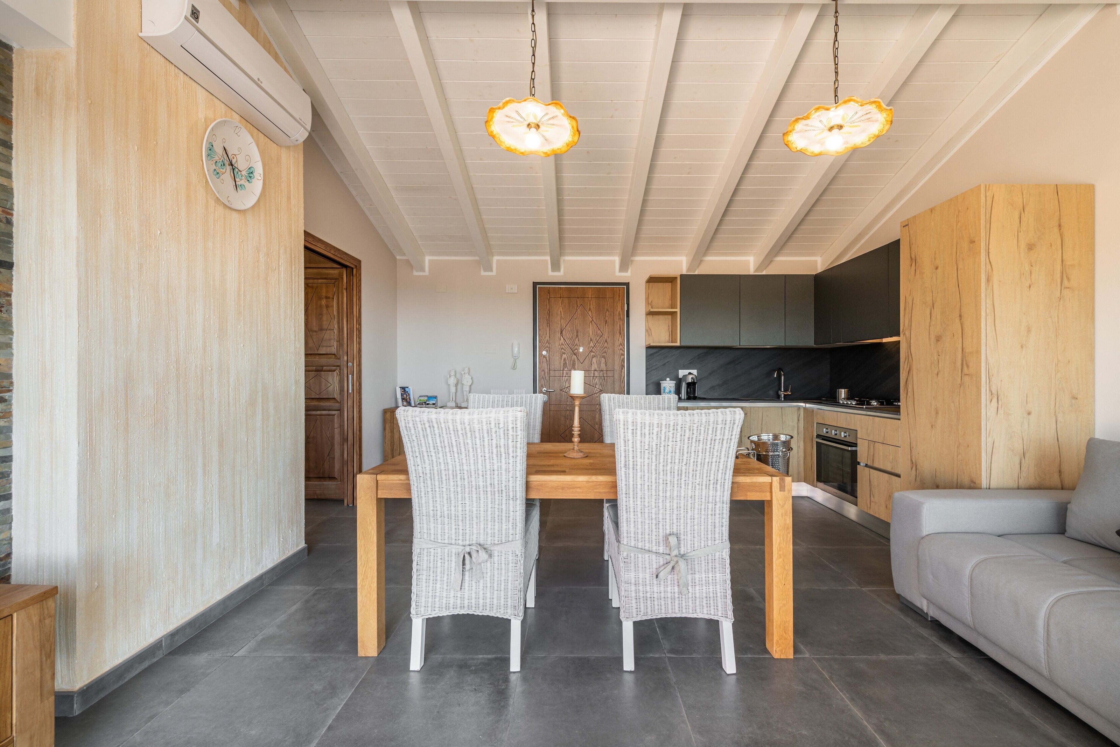 Een eetgedeelte met een houten tafel en rieten stoelen, een keuken met donkere kastjes, en een grijze bank.