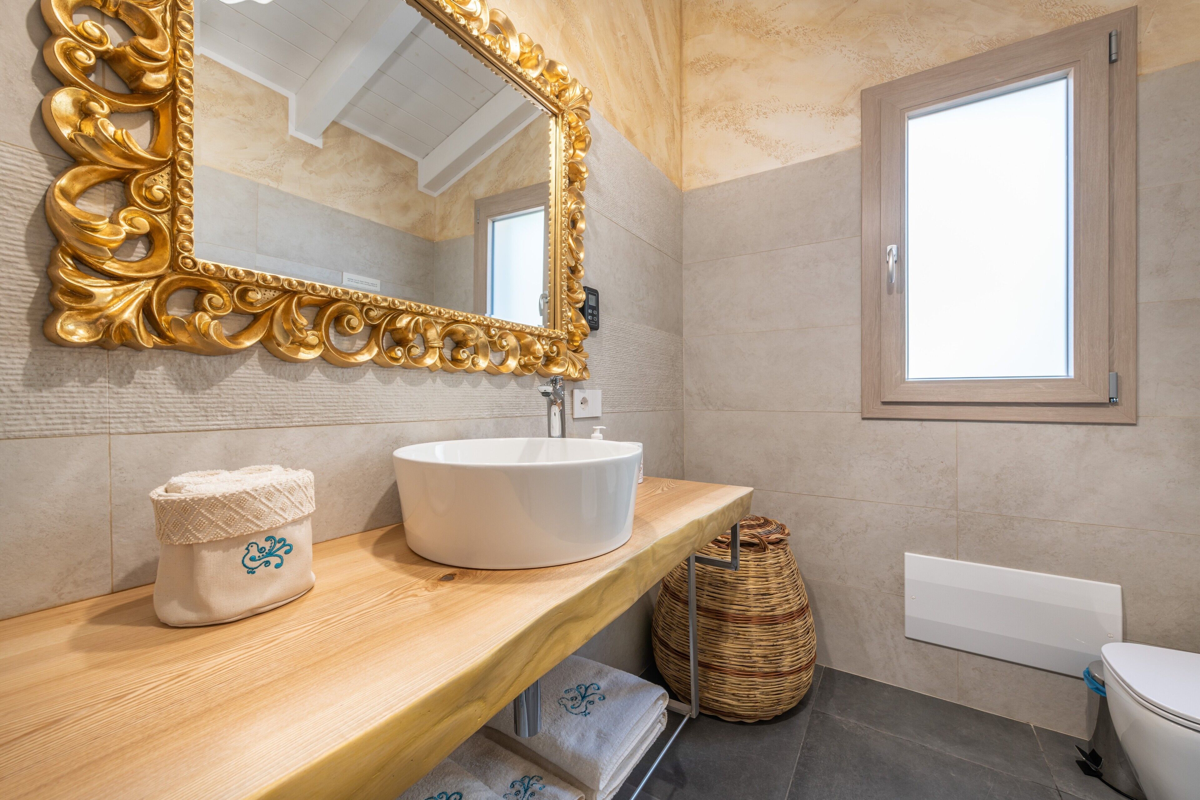 Un baño con un espejo grande y ornamentado, un lavamanos blanco, un tocador de madera, una cesta de mimbre y una ventana.