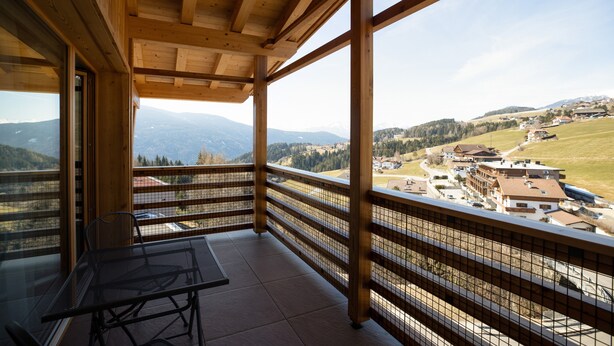 A wooden balcony with a table and chairs, overlooking a mountainous landscape with buildings.