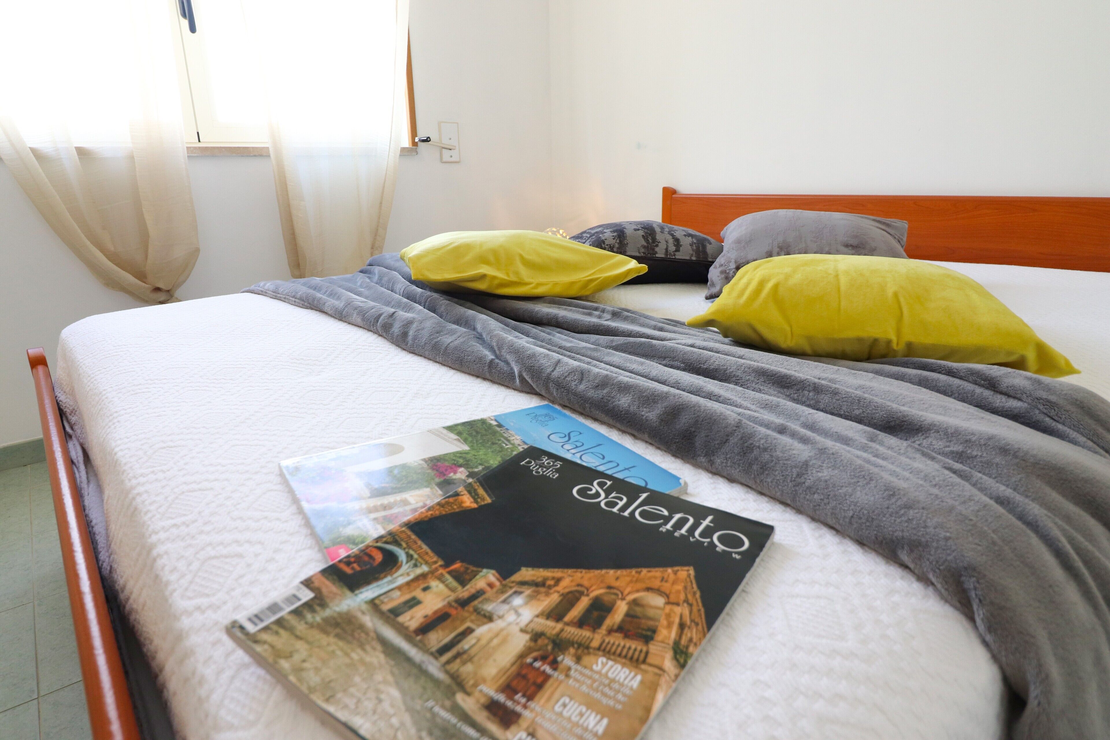 Un lit avec un matelas blanc, des oreillers gris et jaunes, une couverture grise et un magazine intitulé « Salento ».