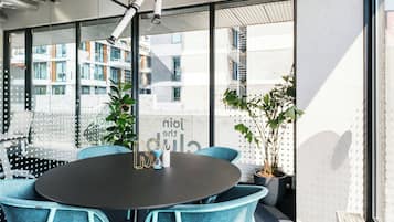 Een moderne vergaderruimte met een ronde tafel, blauwe stoelen en grote ramen met uitzicht op een gebouw met balkons en een trap.