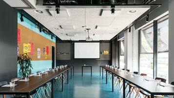 Una sala de conferències amb taules llargues, cadires, una pantalla de projector i un gran finestral.