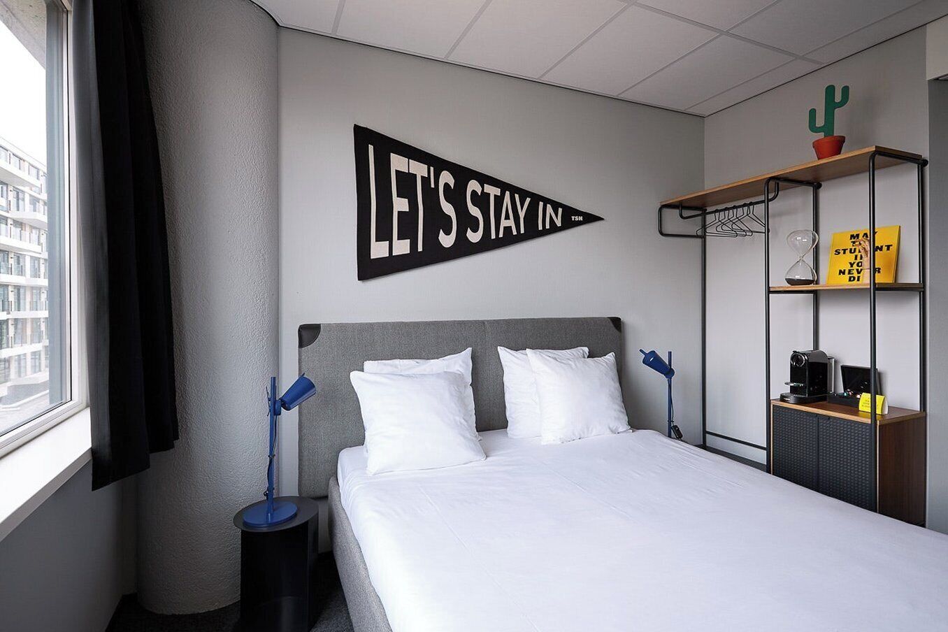 A hotel room with a bed, a wall-mounted sign saying 'LET'S STAY IN,' a window with curtains, a nightstand, a lamp, and a shelf with a cactus and a yellow sign.