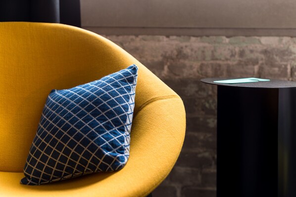 A yellow armchair with a blue checkered pillow, a black side table, and a brick wall in the background.