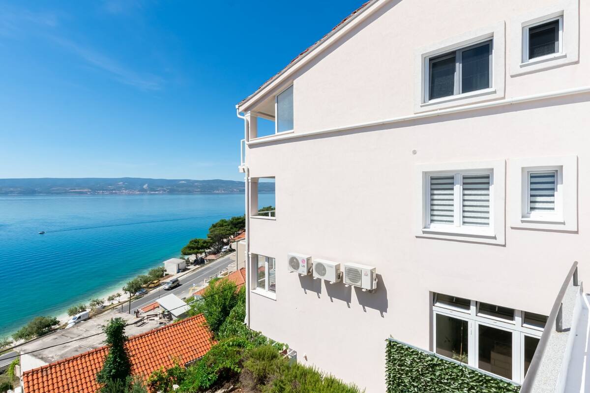 Un bâtiment blanc doté de nombreuses fenêtres et de plusieurs unités de climatisation, qui surplombe une zone côtière avec une mer bleue et limpide et des collines lointaines.