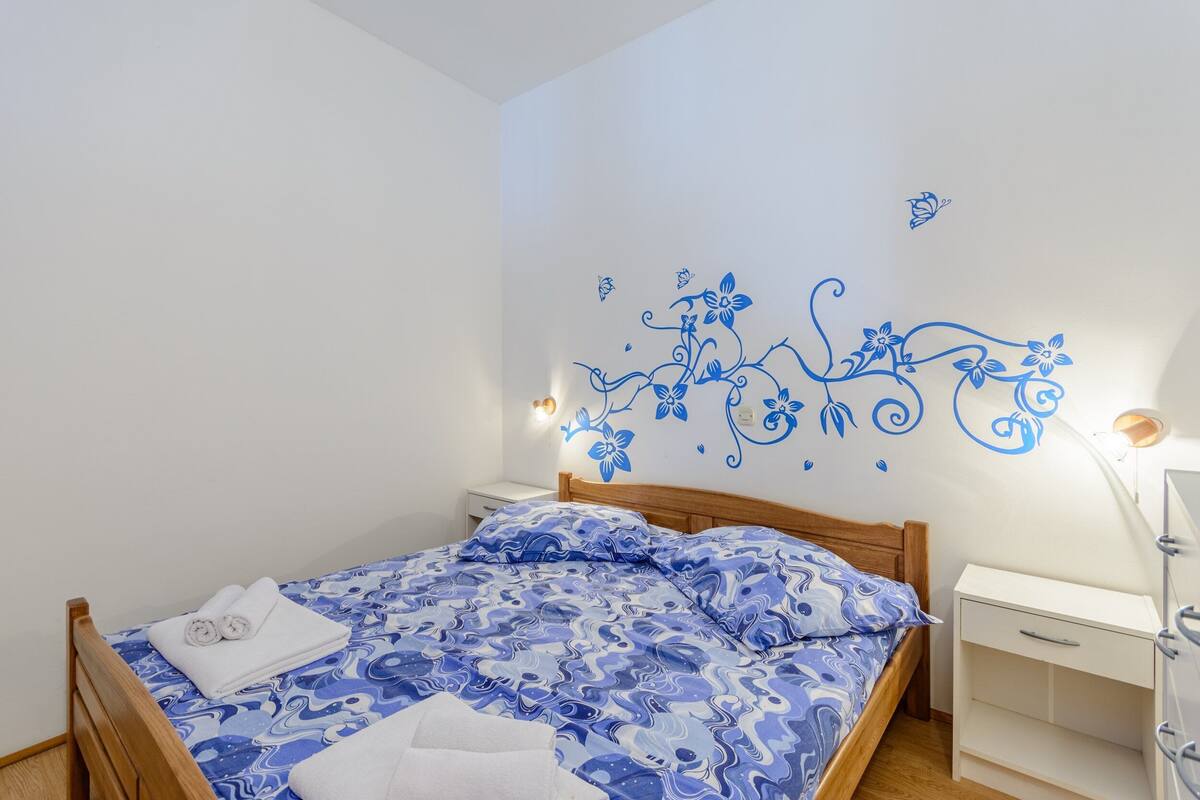 Un lit bien fait, recouvert d’une couvre-lit à motifs bleus et blancs, avec une tête de lit en bois et une table de chevet blanche sur laquelle se trouve une lampe.
