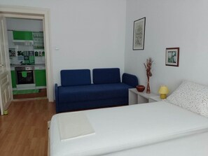 Een kamer met een bed, een blauwe bank, een kleine tafel met een lamp, en een keuken zichtbaar door een geopende deur.