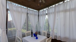 A covered outdoor dining area with white curtains, a table with a bottle and a blue bowl, and chairs. - Apartment Jeanny (Dubrovnik)