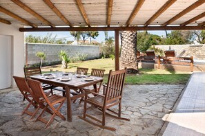 Un patio al aire libre con una mesa y sillas de madera, piso de piedra y vista a un jardín trasero con muro de piedra y follaje.