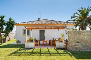 Una casa pequeña con techo de tejas, una pérgola de madera, un patio con sillas y una mesa, y un muro de piedra.