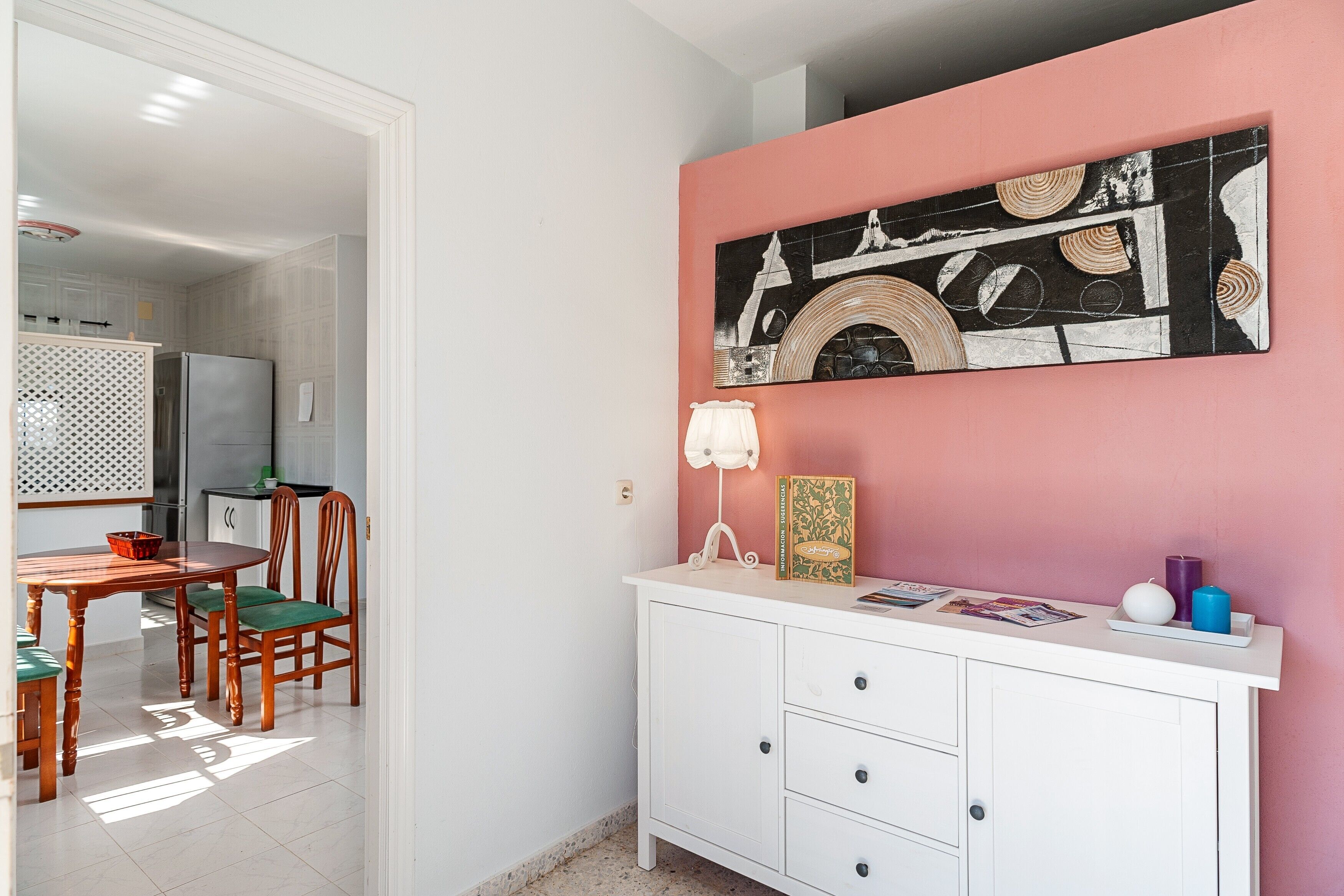 A dining area with a wooden table and chairs, a white cabinet with drawers, and a pink wall with a black and white artwork.