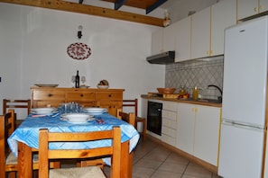 Comedor con mesa preparada para cuatro personas, sillas de madera, cocina con armarios blancos y un refrigerador blanco.