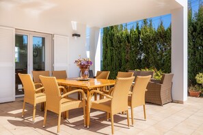 An outdoor patio with a wooden table and wicker chairs, a potted plant, and a tall green hedge.