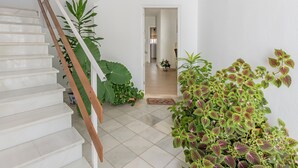 Un escalier aux marches blanches et aux rampes en bois, menant à une pièce dont le sol est recouvert de carreaux et où l'on trouve des plantes en pot.