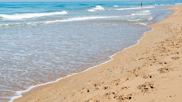 Une plage à l’eau claire, avec un rivage sablonneux, et une personne qui se promène.