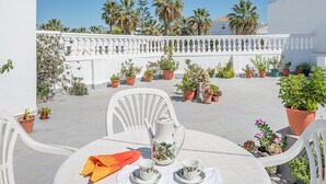 Un espace extérieur aménagé avec des sièges blancs, une table pour deux, entouré de plantes en pot et d’une balustrade blanche.