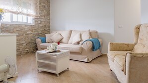 Un salon avec un mur en pierre, un canapé beige, une table d’appoint blanche et un fauteuil à motifs.