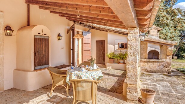 A covered outdoor seating area with wicker chairs, a table with a floral tablecloth, stone walls, wooden beams, and a stone fireplace. - Holiday Home "Son Galiana" with Wi-Fi, Garden and Terrace (Montuïri)
