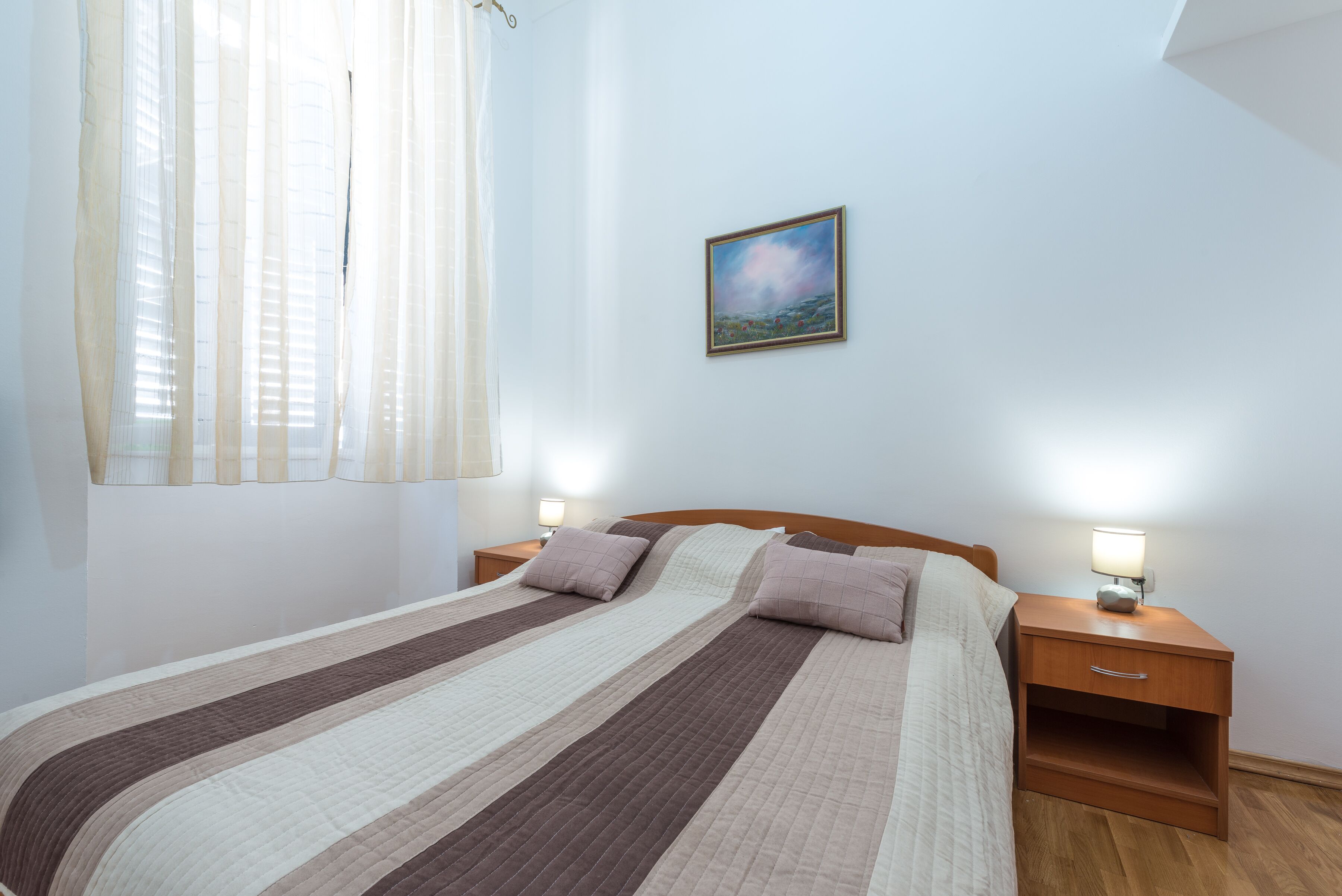Un lit bien fait, recouvert d’une couvre-lit à rayures, deux tables de chevet avec des lampes et un tableau encadré au mur.
