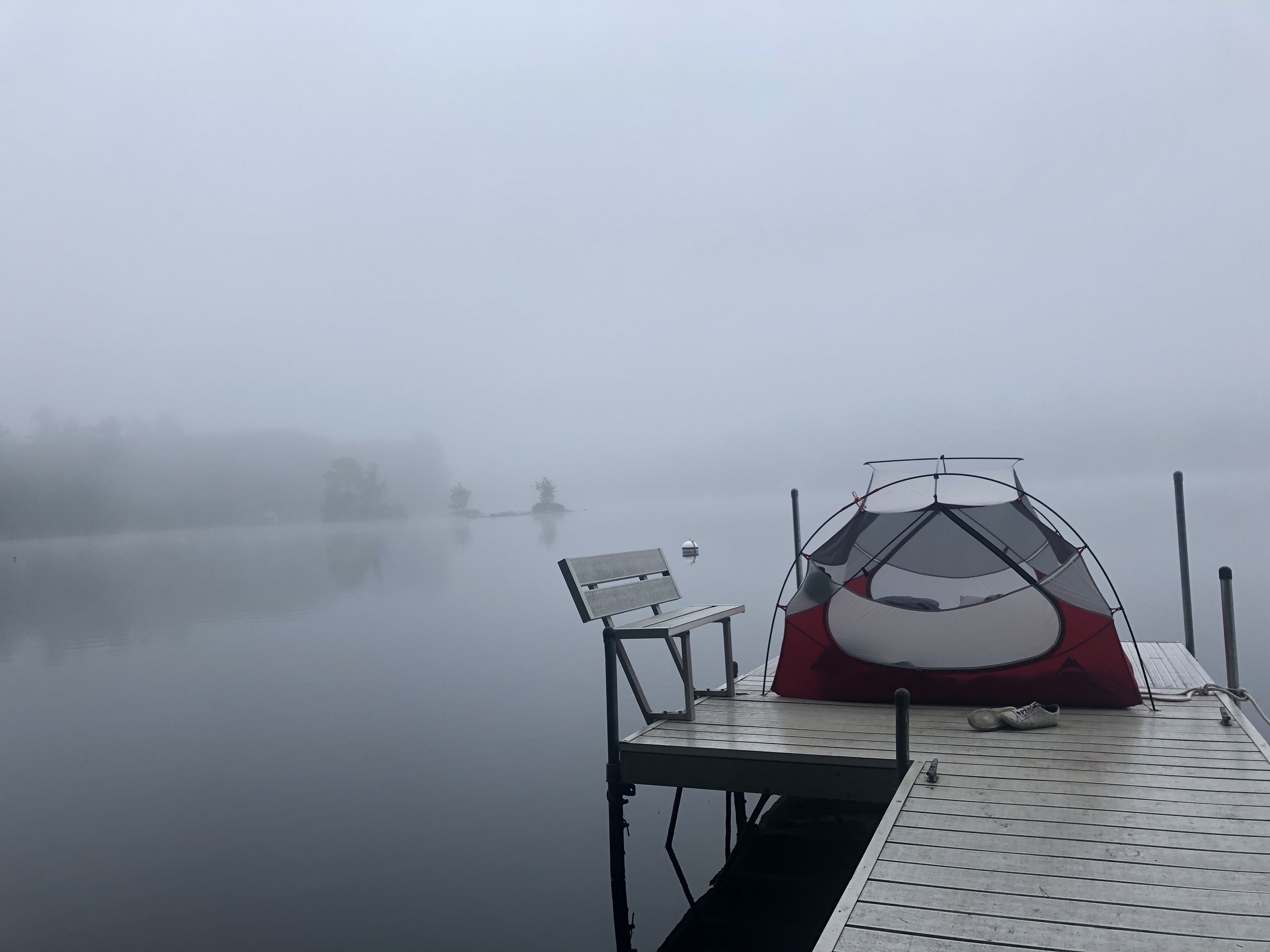 A foggy lakeside scene with a red and white tent and a bench on a dock.