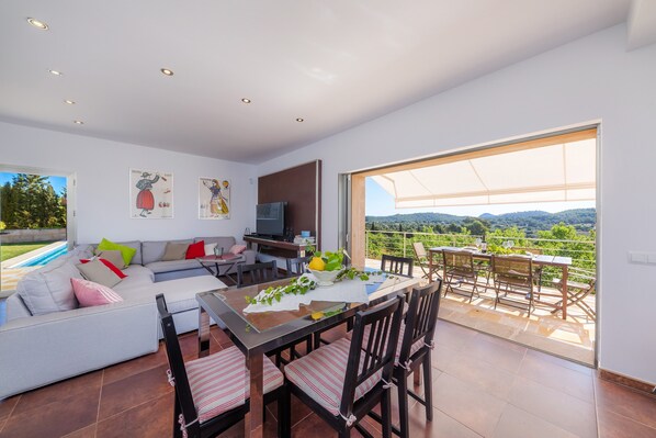A modern living room with a sofa, dining table, and chairs, a TV, and a balcony with a view.