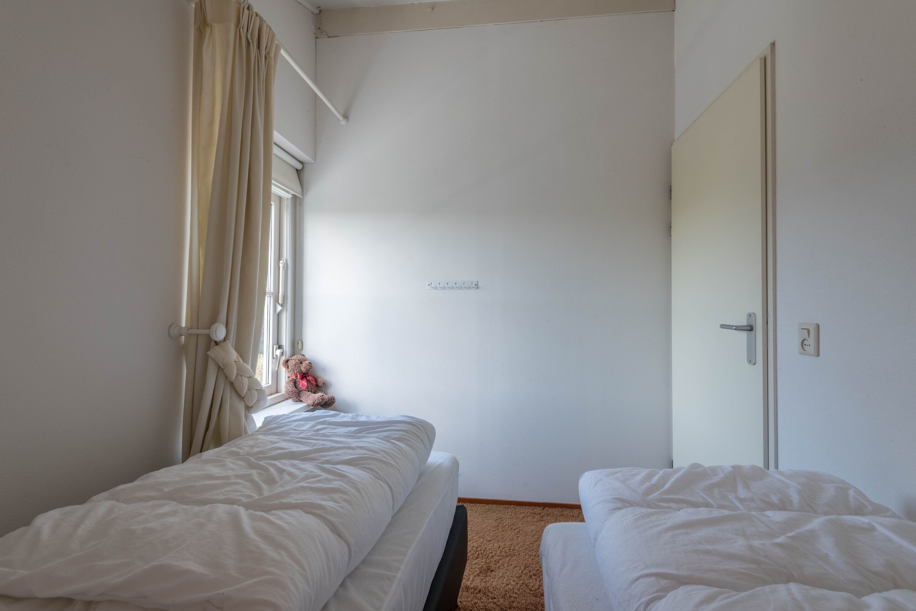 Ein kleiner, sauberer Raum mit zwei Einzelbetten, einem Fenster mit Vorhängen, einer Tür und einer Steckdose an der Wand.