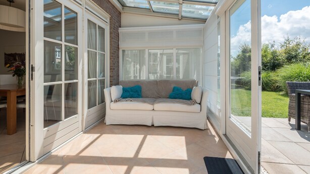 Una sala de estar con techo de cristal, un sofá color beige con cojines azules y vistas a un jardín.