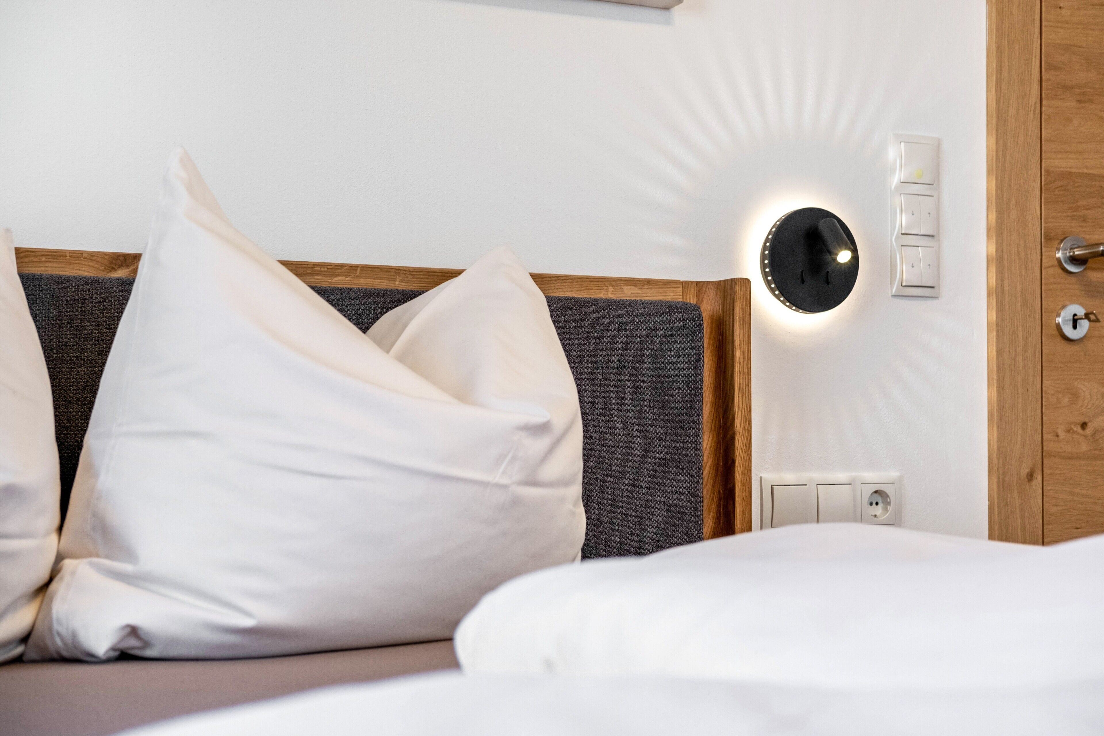 Une chambre d’hôtel avec un lit, des oreillers blancs, une tête de lit, une lampe fixée au mur, une porte en bois et une prise de courant.